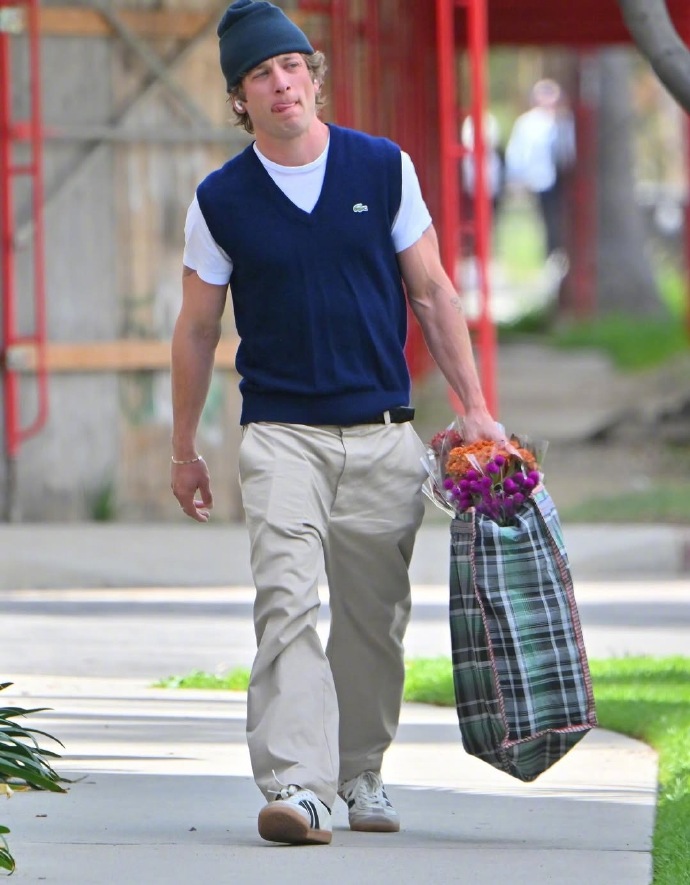 Jeremy Allen White is wearing a vest and carrying a woven bag of fresh flowers.