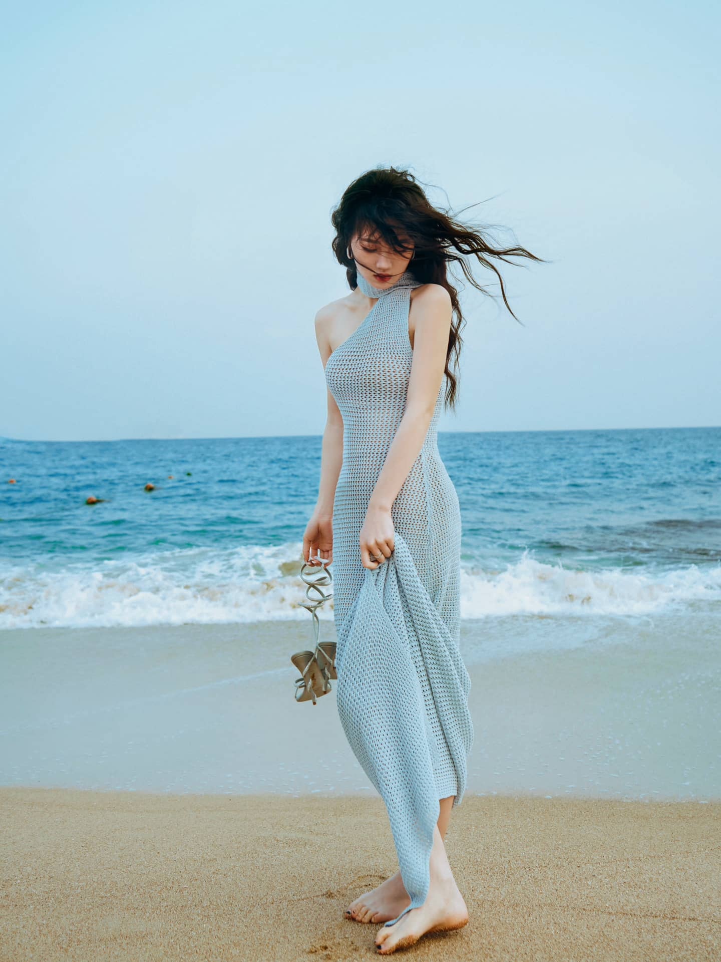 Girl in one-shoulder knitted dress walks barefoot on the beach