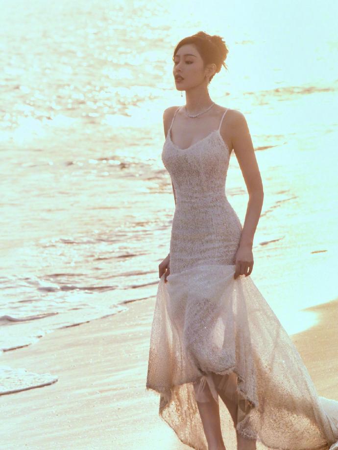 A beautiful woman walks along the seaside at sunset, holding a sheer, spaghetti-strap maxi dress.