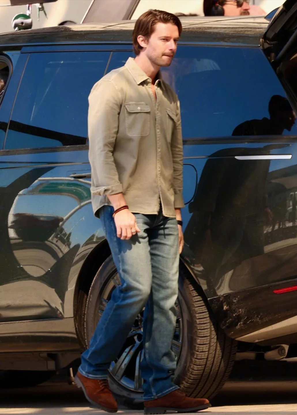 Patrick Schwarzenegger on Set, Wearing a Casual Olive-Green Shirt and Medium-Wash Jeans Outfit with Brown Shoes